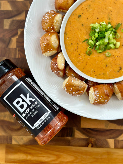Bowl of Black Magic Beer Cheese surrounded by pretzel bites and a jar of Spiceology Black Magic